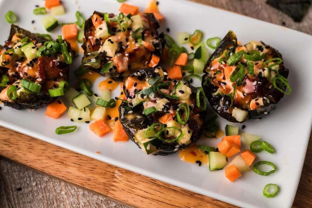 Sushi rolls topped with diced cucumber, carrot, and green onions finished with sesame seeds and a creamy sauce, served stylishly on a white rectangular plate. 