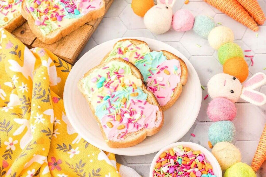 Plate of Unicorn Toast with pastel-colored frosting and sprinkles, surrounded by colorful decorations and a yellow floral cloth.