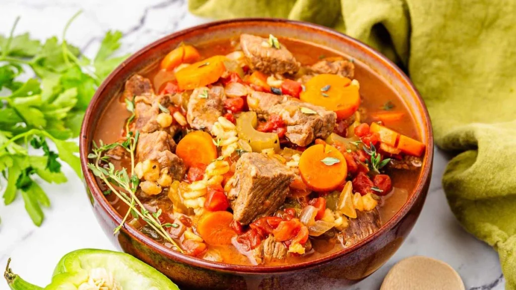 A bowl of vegetable beef stew with chunks of beef, carrots, tomatoes, and herbs in broth.