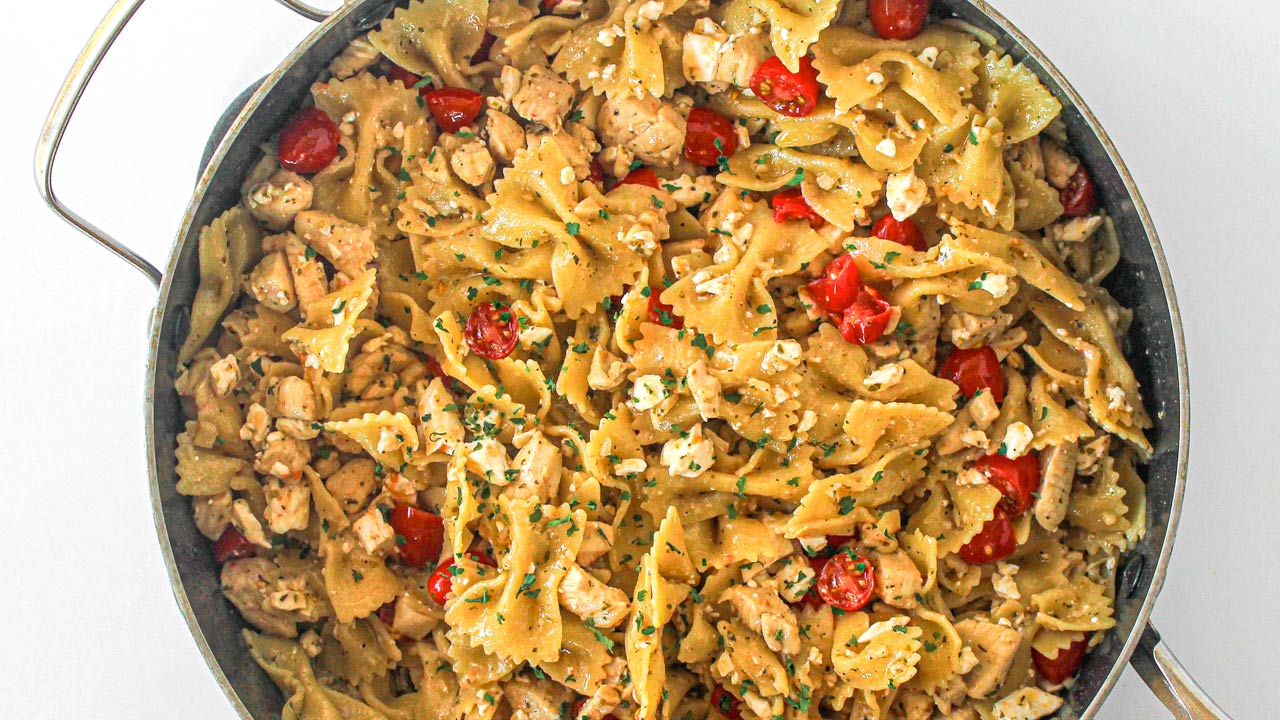A pan filled with bowtie pasta mixed with diced chicken, cherry tomatoes, feta cheese, and chopped herbs.