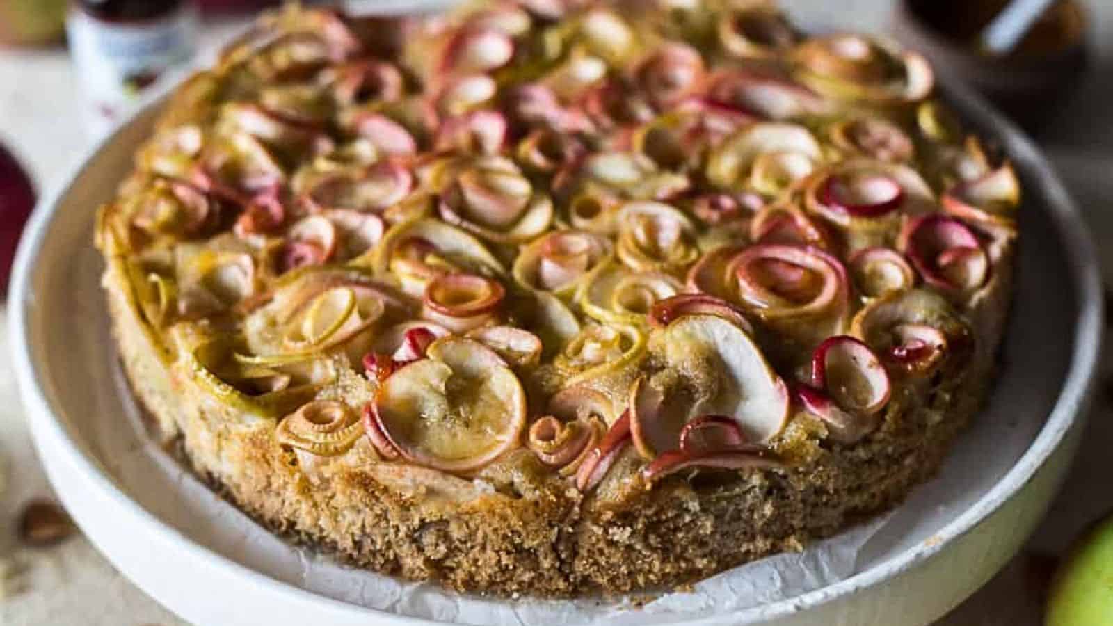 Almond apple cake topped with thinly sliced apples arranged in swirls, on a white cake stand.