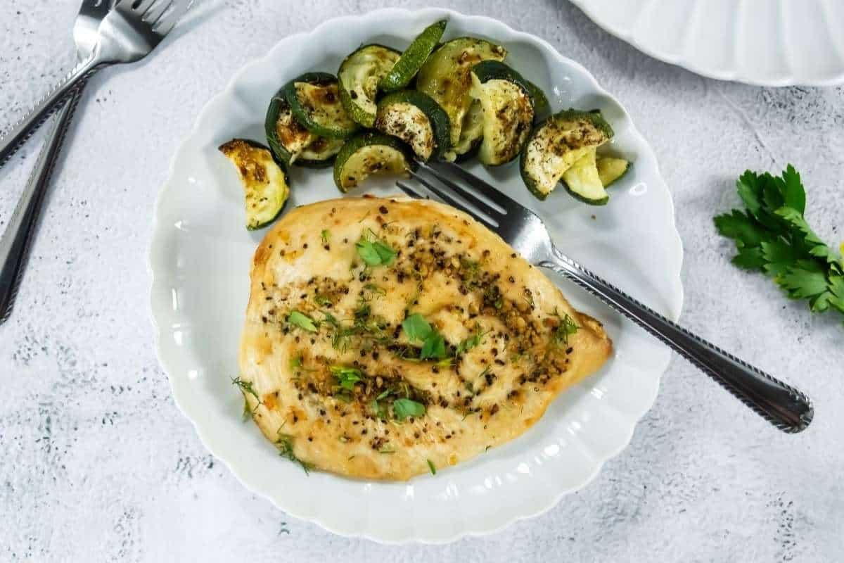 A plate with a broiled chicken breast garnished with herbs and a side of roasted zucchini slices, with a fork and knife on the plate.