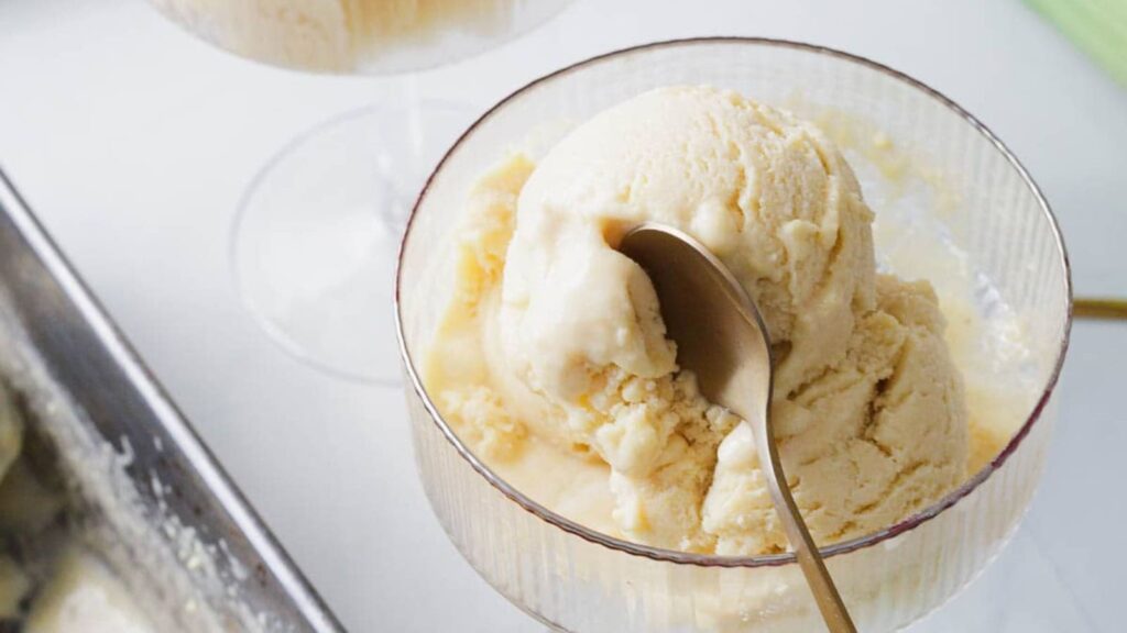 A glass bowl filled with two scoops of vanilla ice cream, with a gold spoon resting in the ice cream. Another bowl and a metal container with more ice cream are visible in the background.