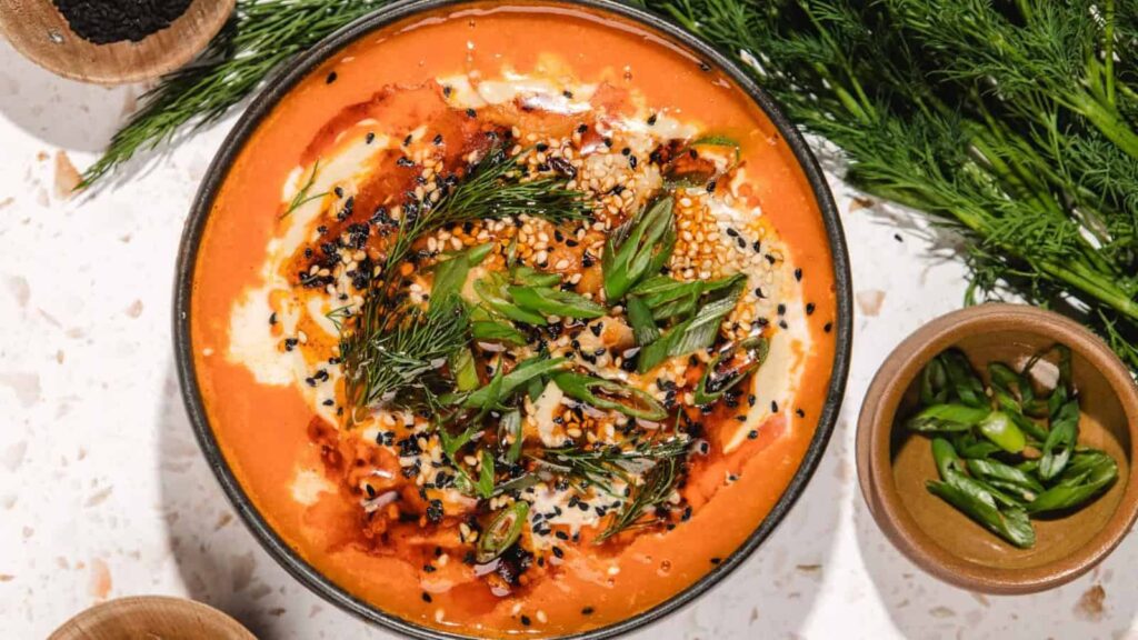 Bowl of creamy soup topped with herbs, seeds, and spices, with fresh dill and green onions on the side.