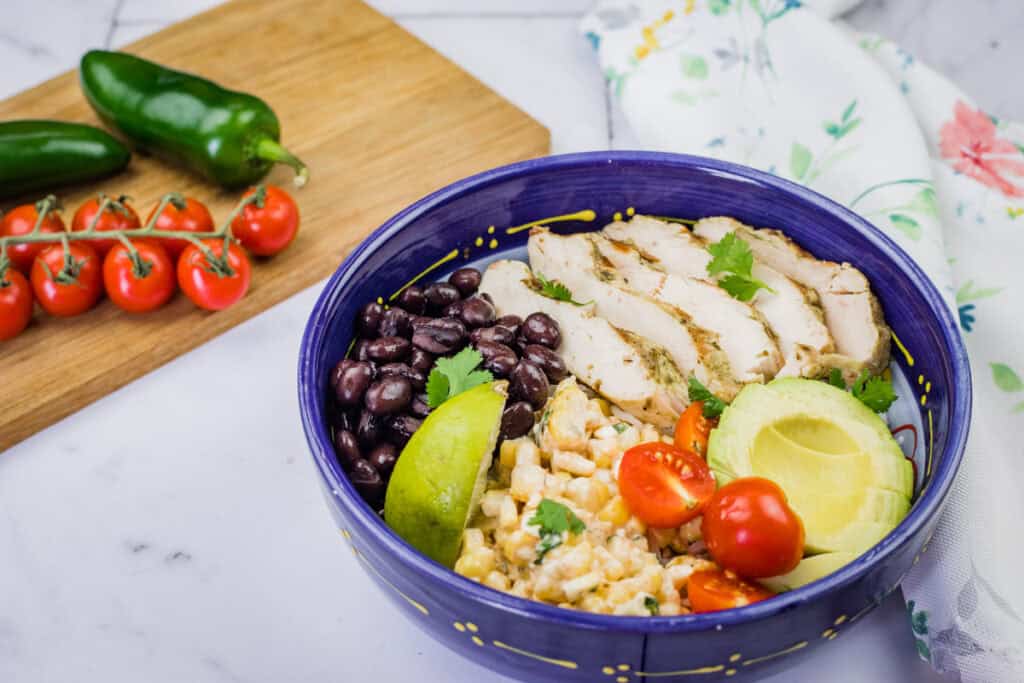 A blue bowl filled with street corn chicken rice bowl, featuring grilled chicken, corn, tomatoes, avocado, and black beans.