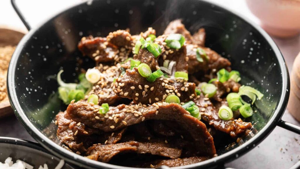 A bowl of cooked beef slices topped with sesame seeds and chopped green onions.
