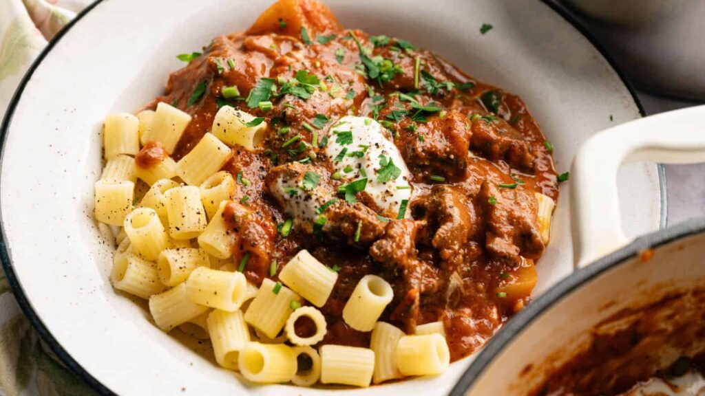 A plate of pasta with beef stew in tomato sauce, topped with a dollop of sour cream and chopped herbs.