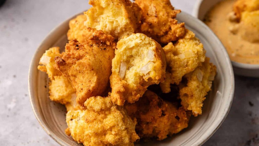 A bowl filled with golden, crispy hush puppies, some with visible onions, sits next to a small dish of dipping sauce.