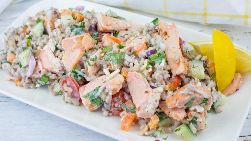 A rectangular white plate of Mediterranean Salmon Salad with Barley, topped with fresh herbs, and a lemon wedge on the side.