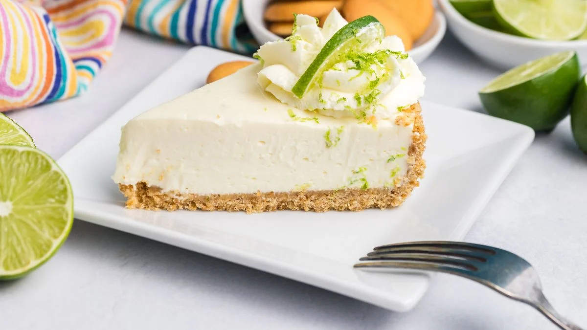Slice of key lime pie with whipped cream and lime zest on a white plate, cookies and limes in the background.