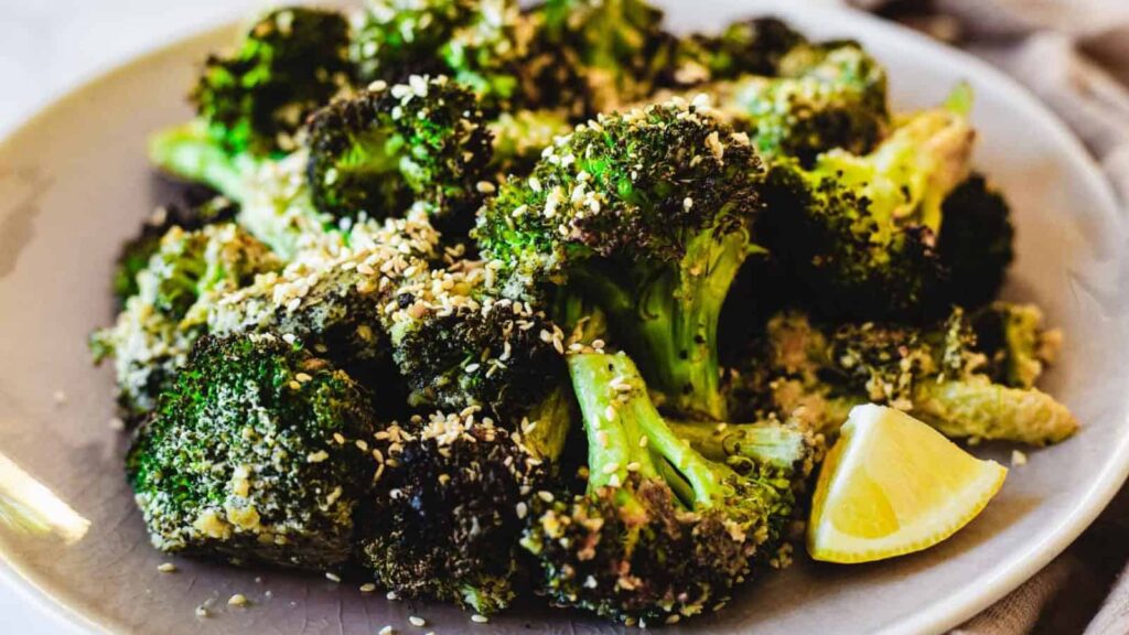 Roasted broccoli topped with sesame seeds on a plate, served with lemon wedges.