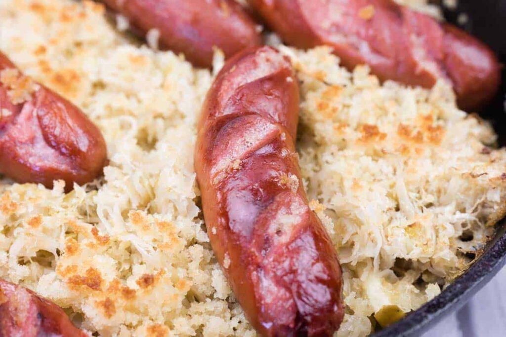 Close-up of browned sausages on a bed of sauerkraut and breadcrumbs in a baking dish.