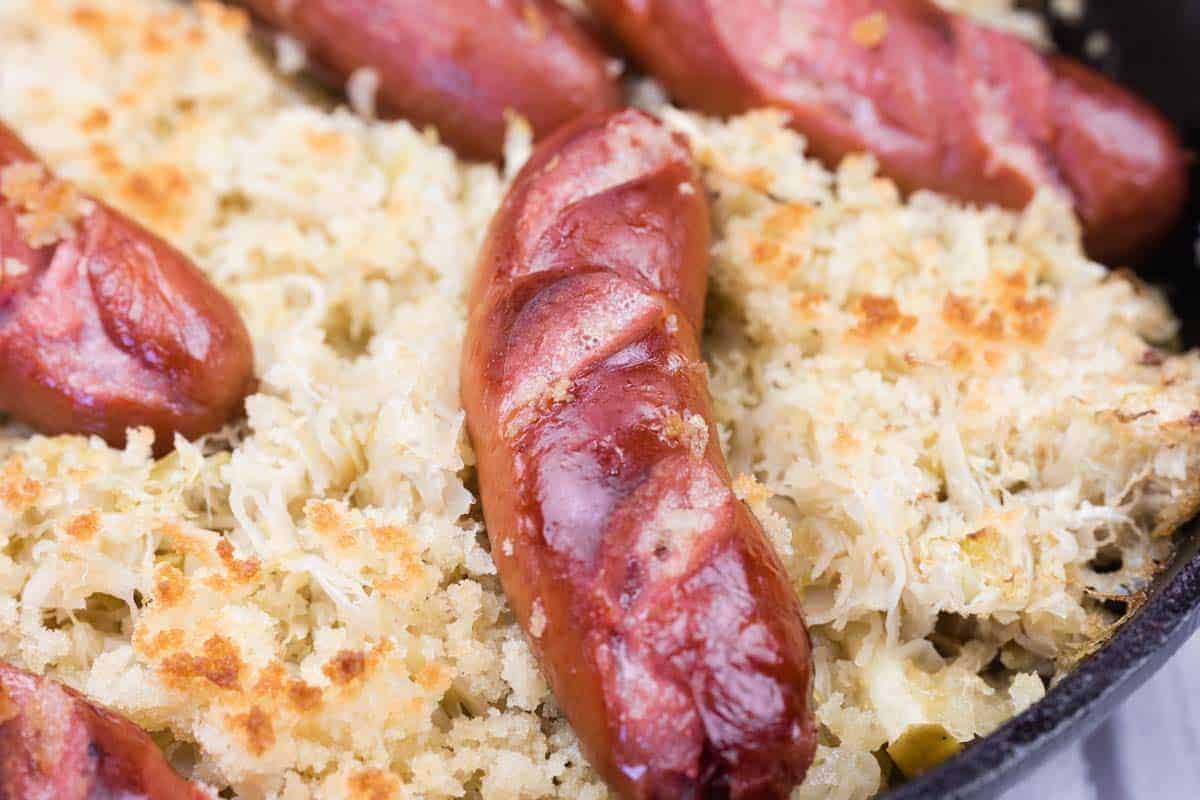 Close-up of browned sausages on a bed of sauerkraut and breadcrumbs in a baking dish.