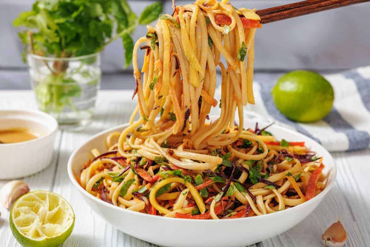 A bowl of Thai Noodle Salad with Mango and Spicy Peanut Dressing is lifted by chopsticks.
