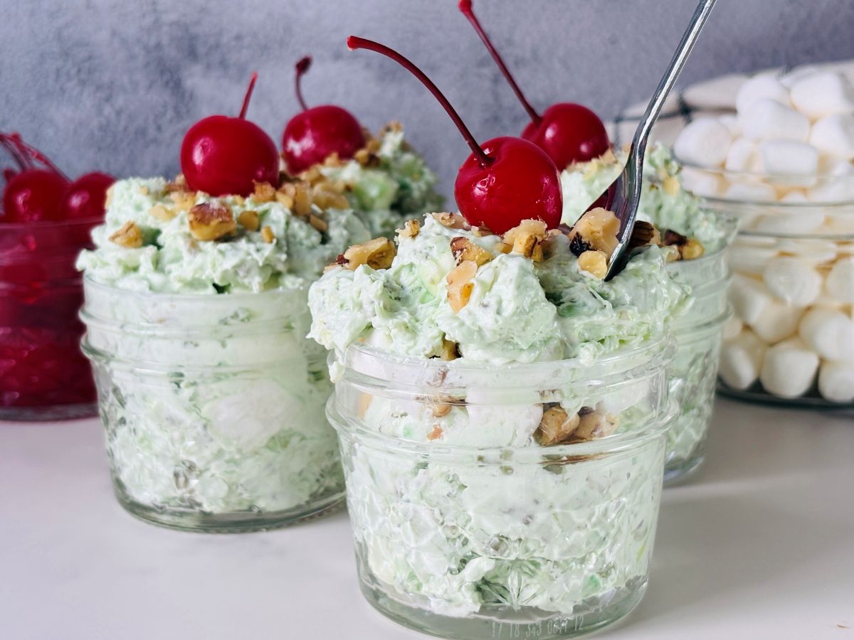 Three glass jars filled with green pistachio salad topped with chopped nuts and maraschino cherries, with mini marshmallows in the background.