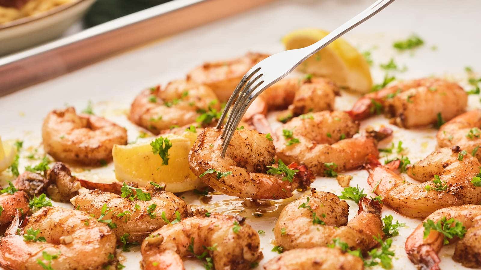 A fork lifting a juicy, baked shrimp from a tray garnished with fresh parsley and lemon wedges.