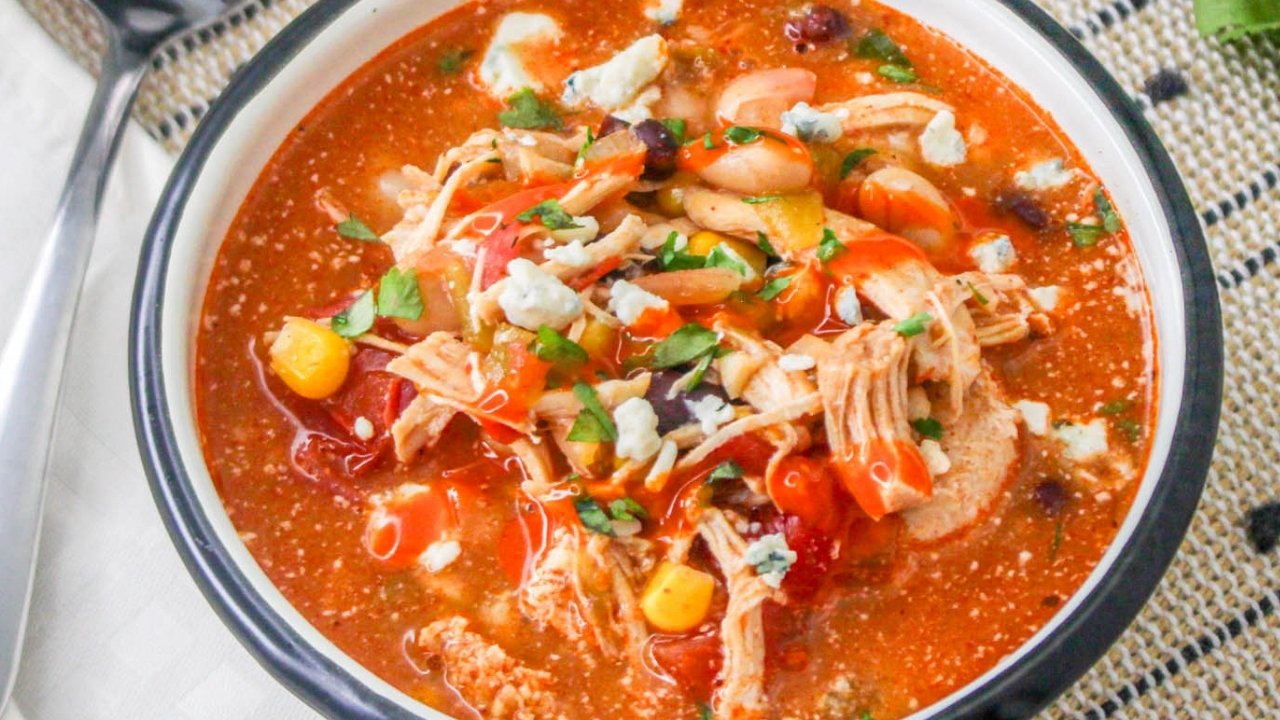 A bowl of Slow Cooker Buffalo Chicken Chili filled with shredded chicken, black and white beans, corn, diced tomatoes, and topped with blue cheese crumbles, chopped parsley, and a drizzle of buffalo sauce.