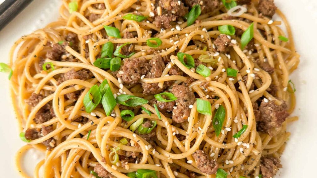 A plate of Chinese Mongolian beef noodles garnished with fresh green onions and roasted sesame seeds.