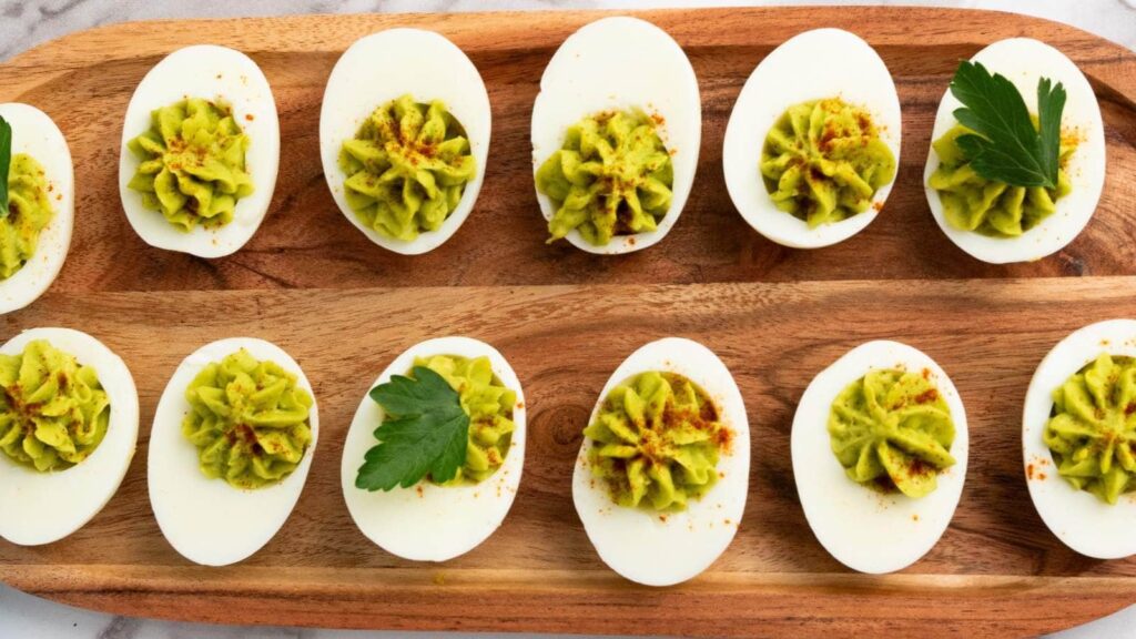 Deviled eggs topped with green filling and paprika are arranged on a wooden serving tray, garnished with parsley leaves.