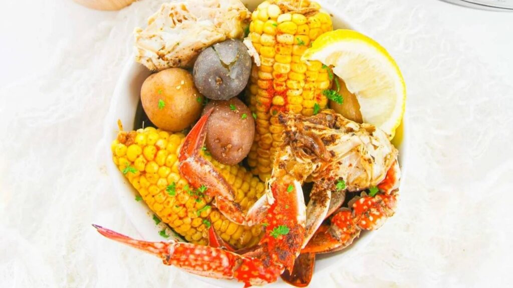 A bowl containing crab legs, corn on the cob, small potatoes, a lemon wedge, and sprinkled herbs.