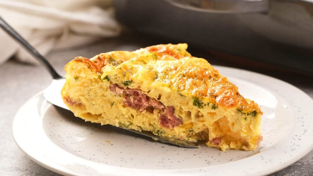 A slice of baked omelet with visible pieces of ham and herbs sits on a spatula over a white plate.