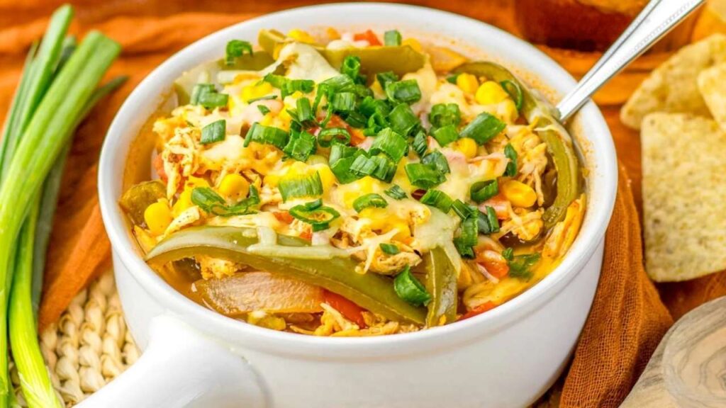 A white bowl filled with shredded chicken, corn, sliced green peppers, melted cheese, and chopped green onions, with a spoon inside.