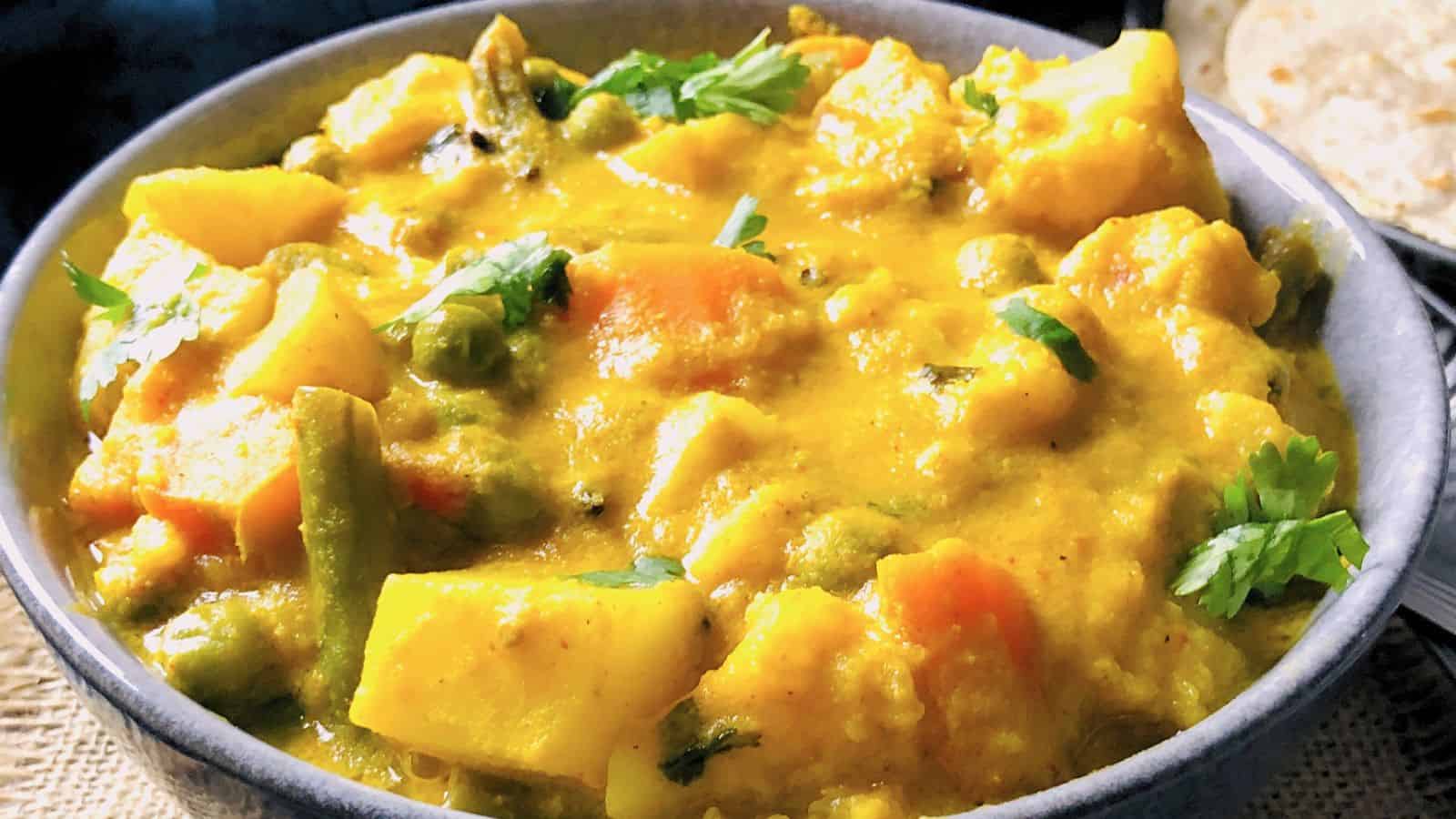 A bowl of colorful mixed vegetable curry garnished with fresh cilantro, served with flatbread on the side.
