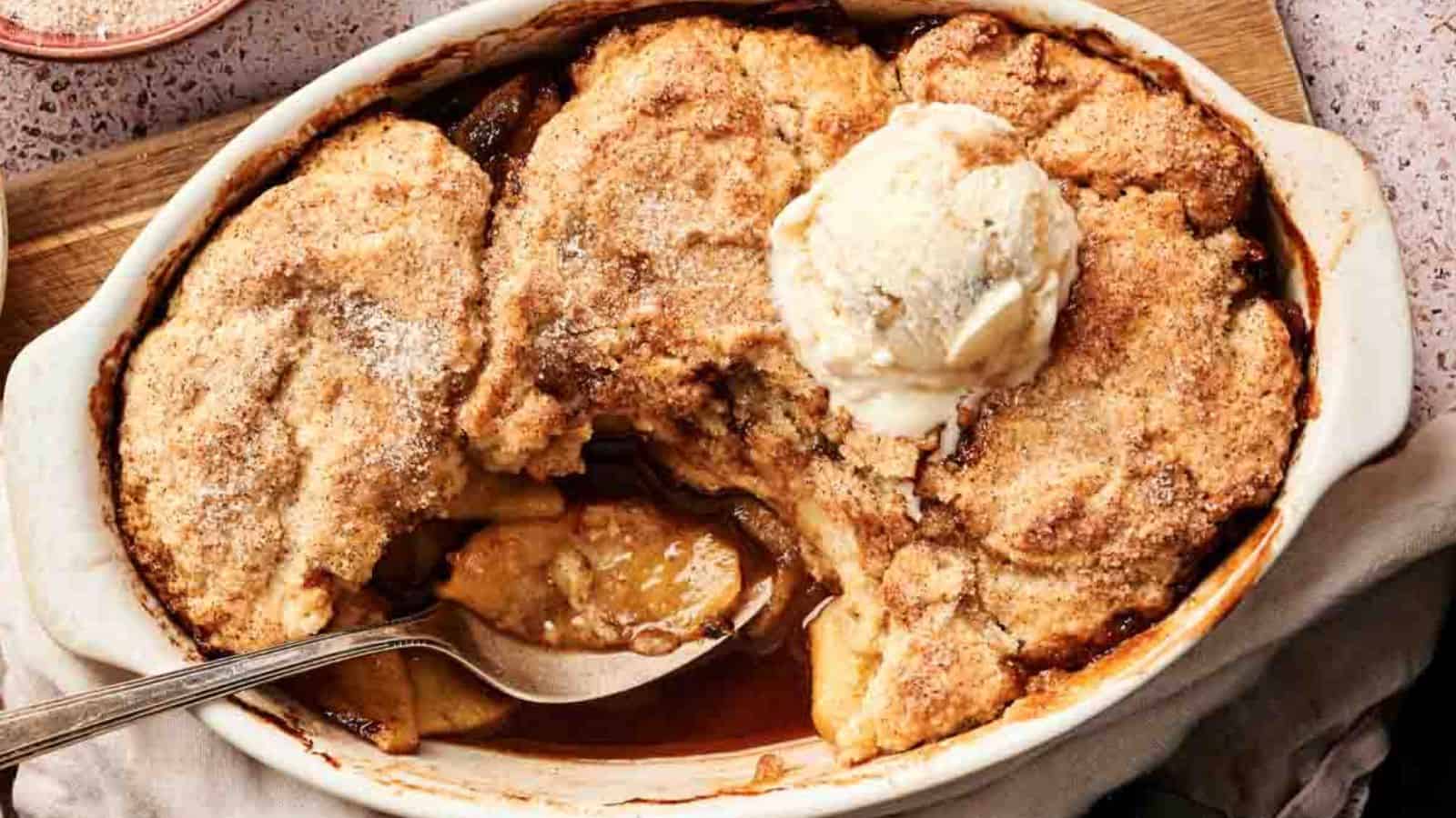 A baking dish filled with apple cobbler topped with a scoop of vanilla ice cream; a serving has been scooped out with a spoon.