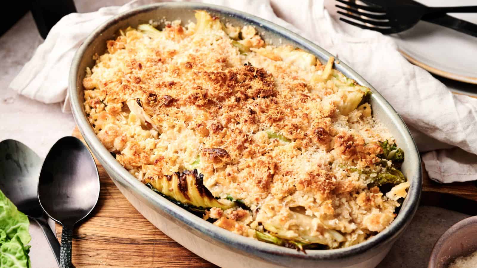 Oval baking dish with a golden-brown breadcrumb topping over a baked casserole, set on a wooden board with serving utensils and plates nearby.