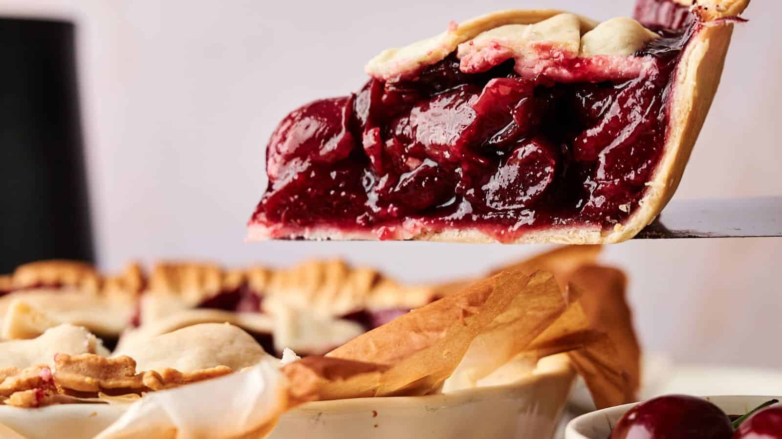 A slice of cherry pie with thick fruit filling is being lifted from a pie dish.