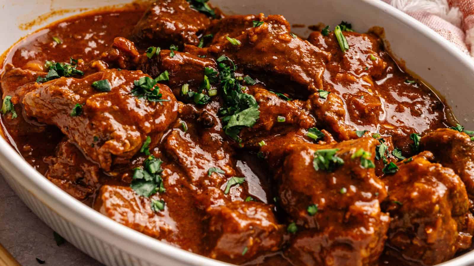 A close-up of beef stew in a thick, red sauce, garnished with chopped fresh herbs, served in a white dish.