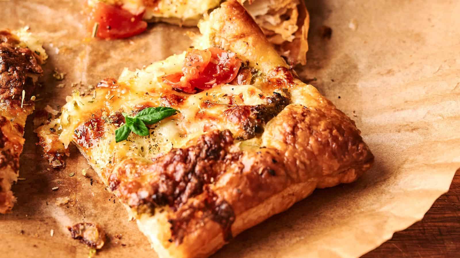 A slice of puff pastry topped with melted cheese, tomatoes, herbs, and a basil leaf, resting on a sheet of brown parchment paper.