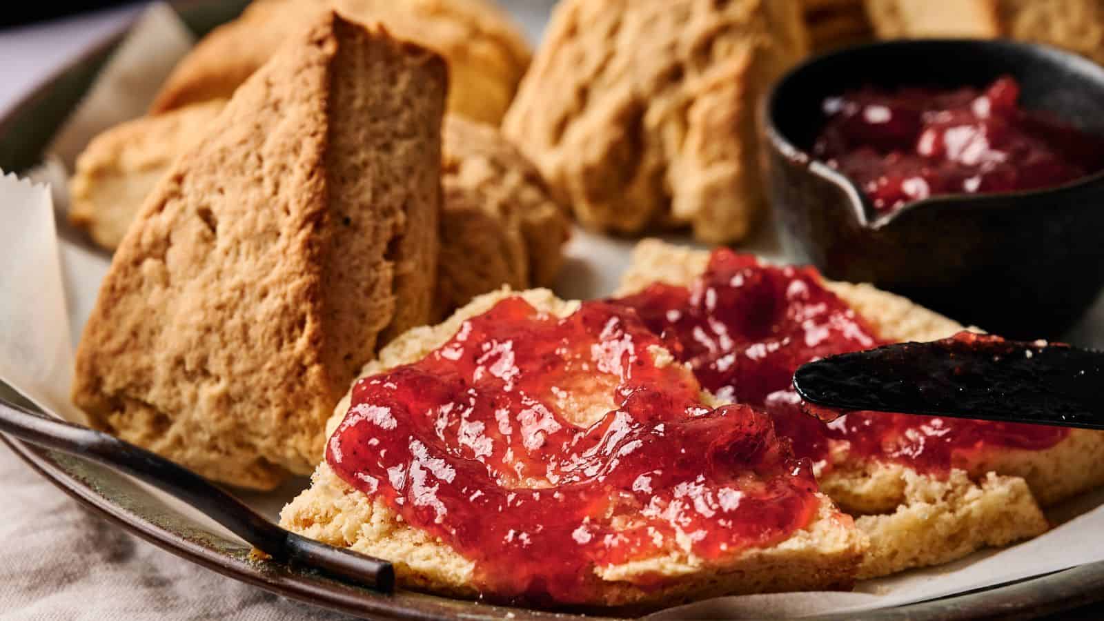 A plate of scones, some whole and some split open, with one scone spread with red jam using a knife. A small bowl of jam is also visible.