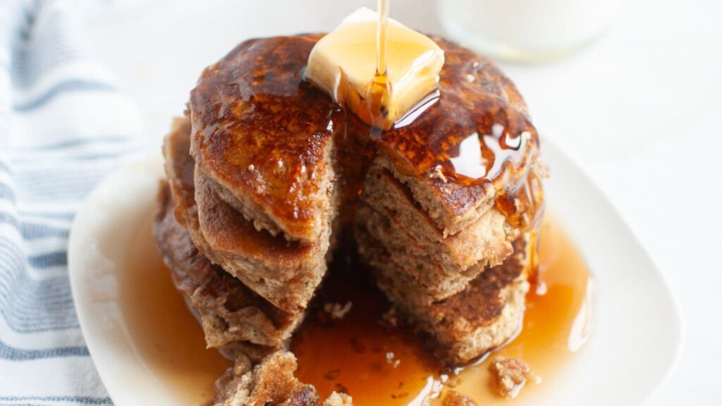 A stack of pancakes with a pat of butter on top is drizzled with syrup on a white plate. A glass of milk and a striped napkin are in the background.