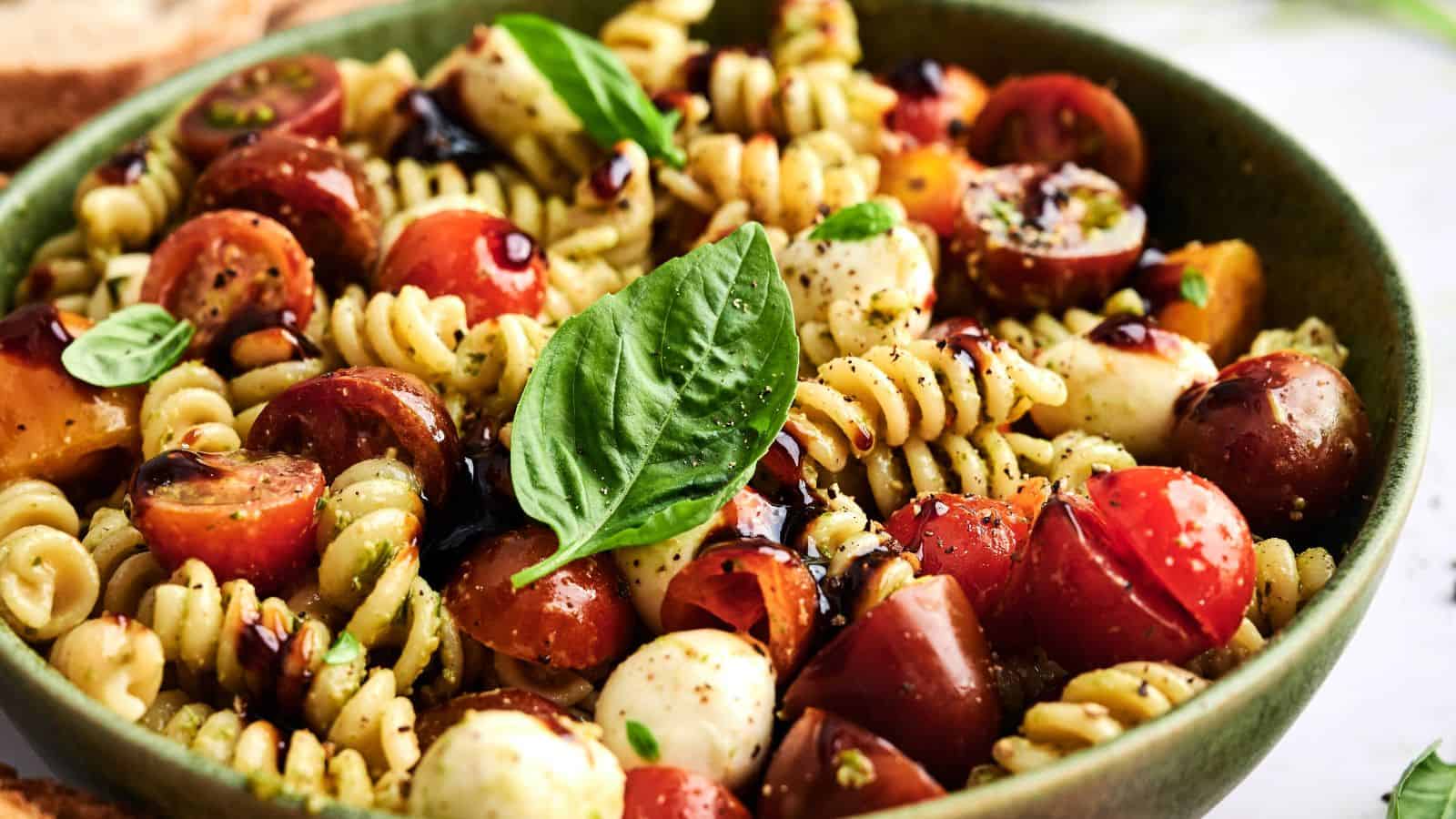 A bowl of rotini pasta salad with cherry tomatoes, mozzarella balls, fresh basil leaves, and a drizzle of balsamic glaze.
