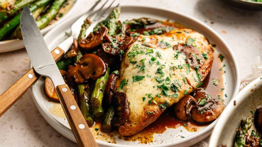 A plate of chicken breast topped with melted cheese and herbs, served with sautéed mushrooms and asparagus, with a knife and fork on the side.