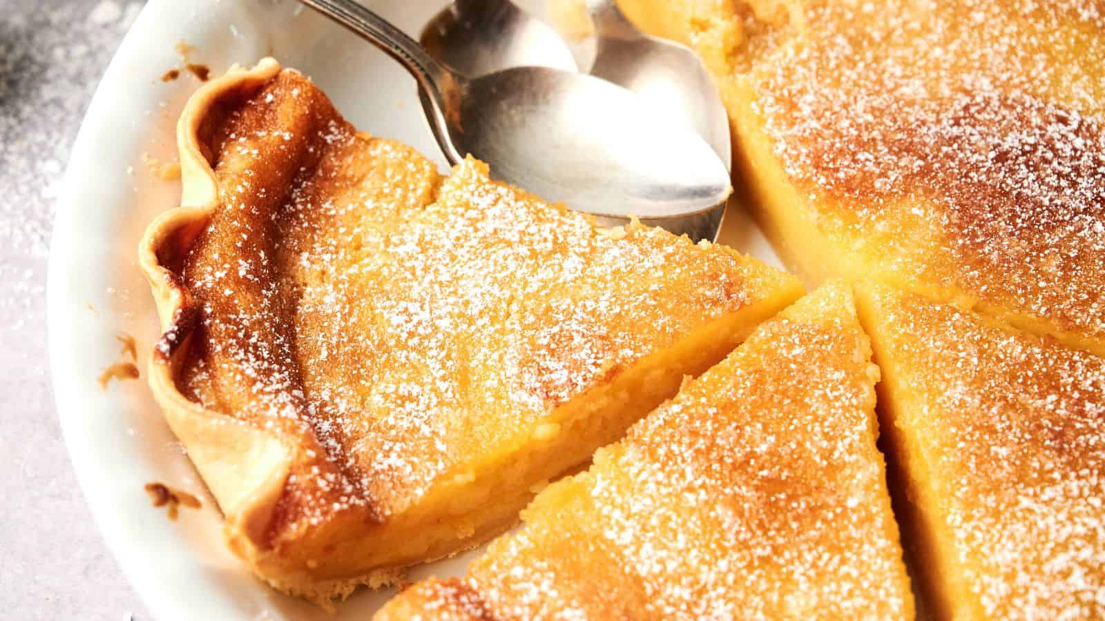 A close-up of a sliced custard pie dusted with powdered sugar, with two metal spoons resting on top of the pie in a white dish.