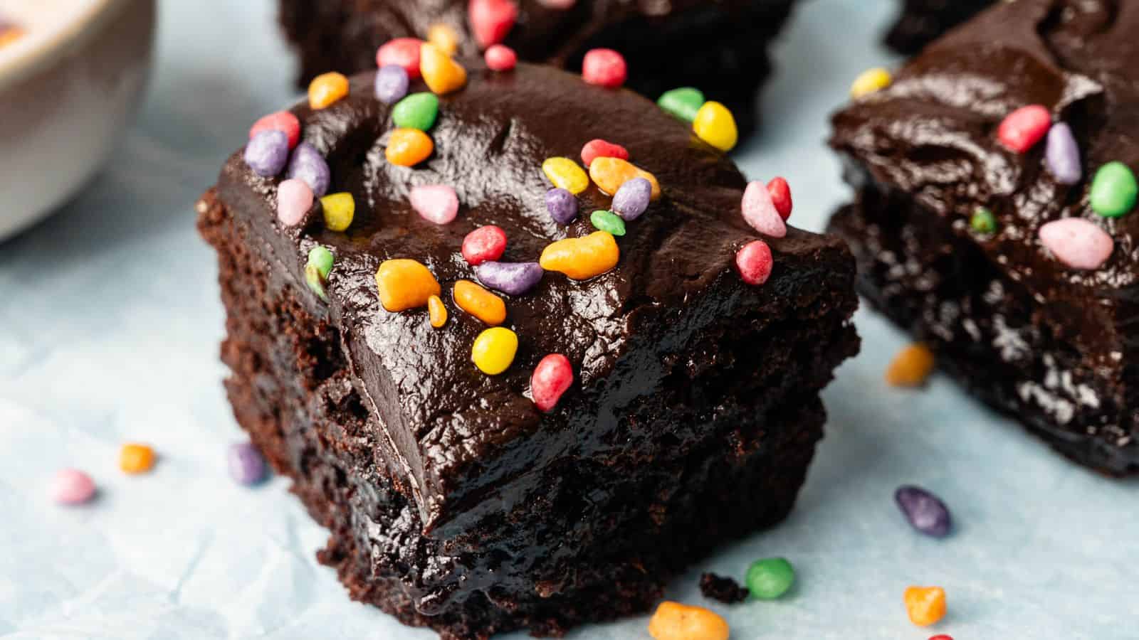 Close-up of a thick, fudgy chocolate brownie topped with colorful candy sprinkles on a light blue surface.