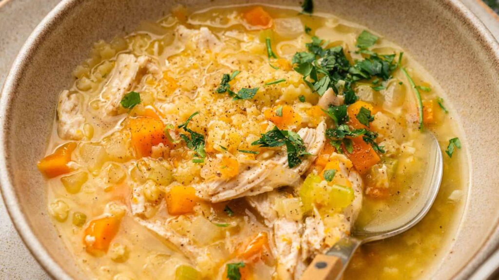 A bowl of chicken and rice soup with chunks of chicken, diced carrots, celery, and parsley garnish, served with a spoon.