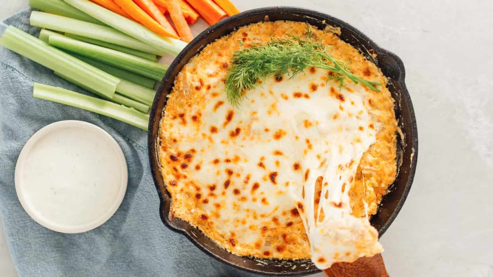 A skillet with melted cheese dip, garnished with dill. Celery and carrot sticks are on the side, next to a bowl of dipping sauce.