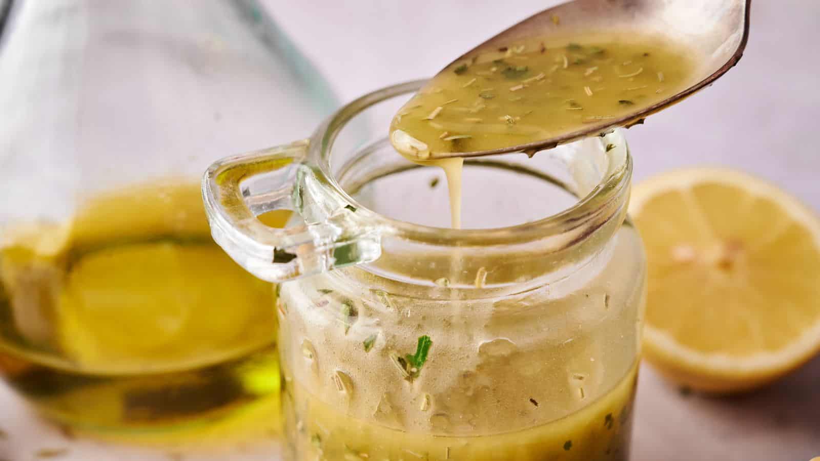 A spoon drizzles vinaigrette from a glass jar, with herbs visible in the dressing; a halved lemon and a bottle of oil are in the background.