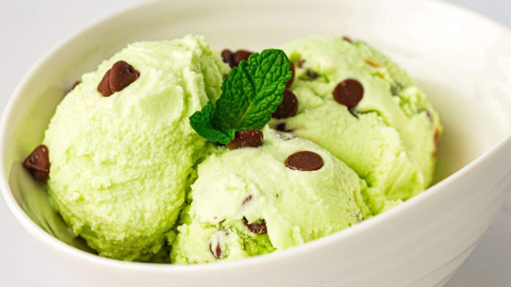 Three scoops of mint chocolate chip ice cream with chocolate chips, garnished with a sprig of mint, in a white bowl.