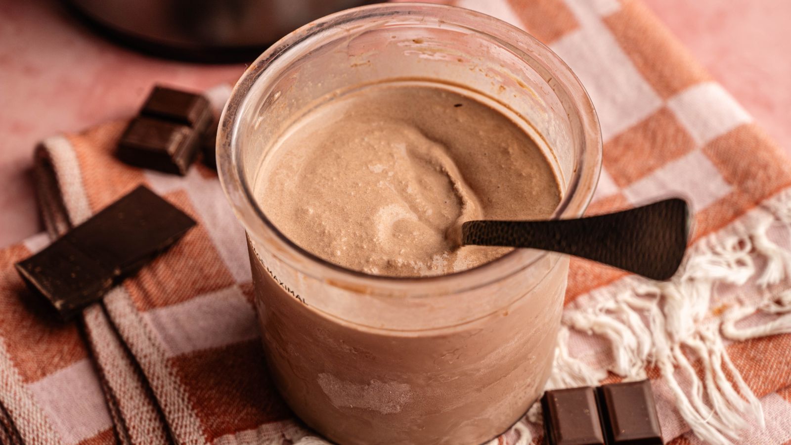 A glass of chocolate milkshake with a spoon inside, placed on a checkered cloth next to pieces of chocolate.