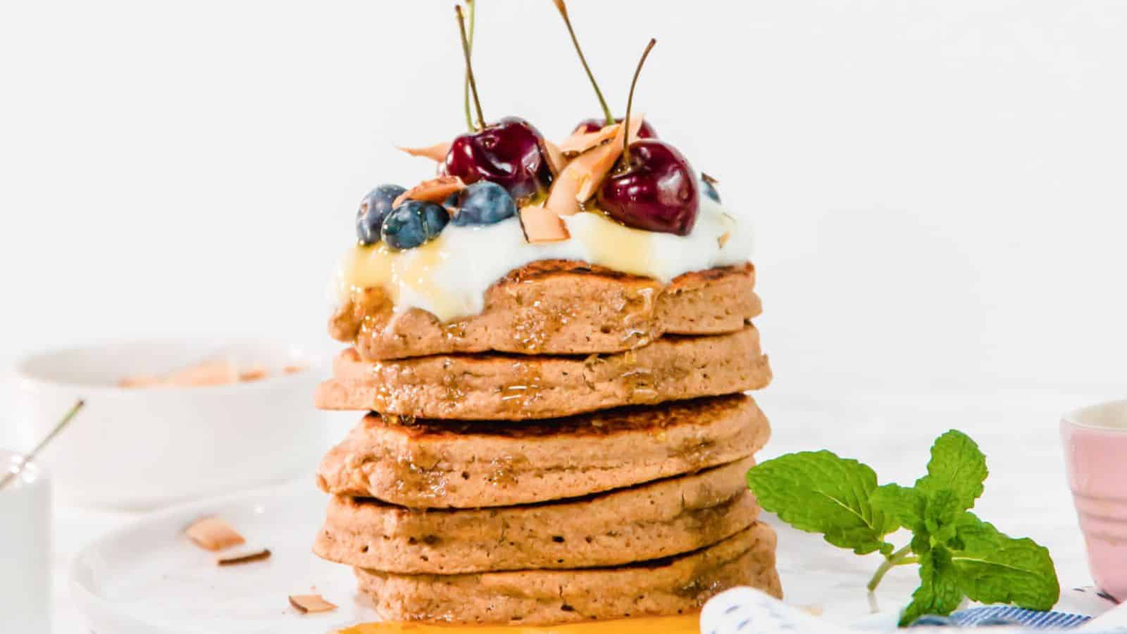 A stack of pancakes topped with yogurt, cherries, blueberries, and coconut flakes, garnished with mint leaves.