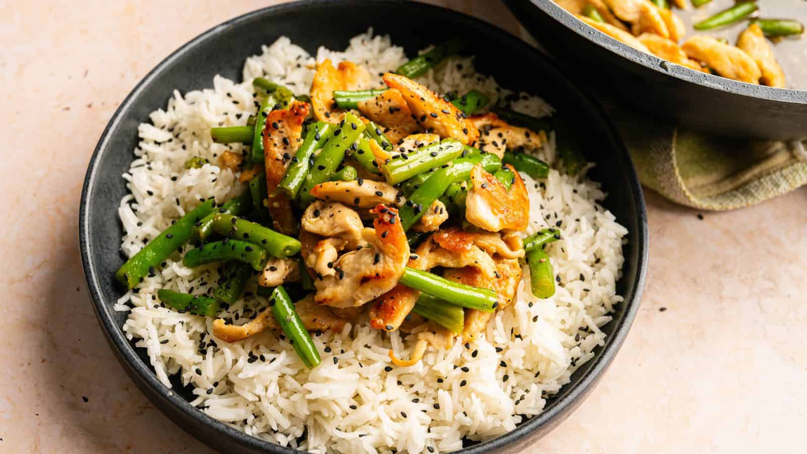 A bowl of white rice topped with stir-fried chicken, green beans, and carrots, garnished with black sesame seeds.