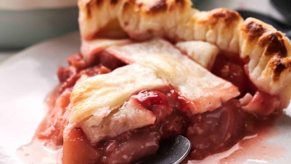 Close-up of a slice of fruit pie with a golden, flaky crust. The filling is a mix of pink and red fruit, slightly oozing out onto the plate.