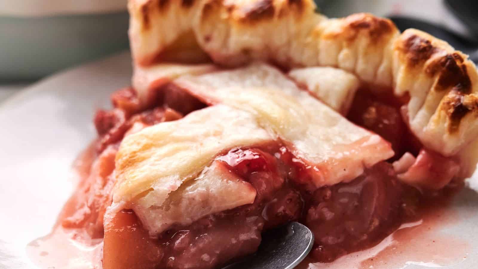 Close-up of a slice of fruit pie with a golden, flaky crust. The filling is a mix of pink and red fruit, slightly oozing out onto the plate.