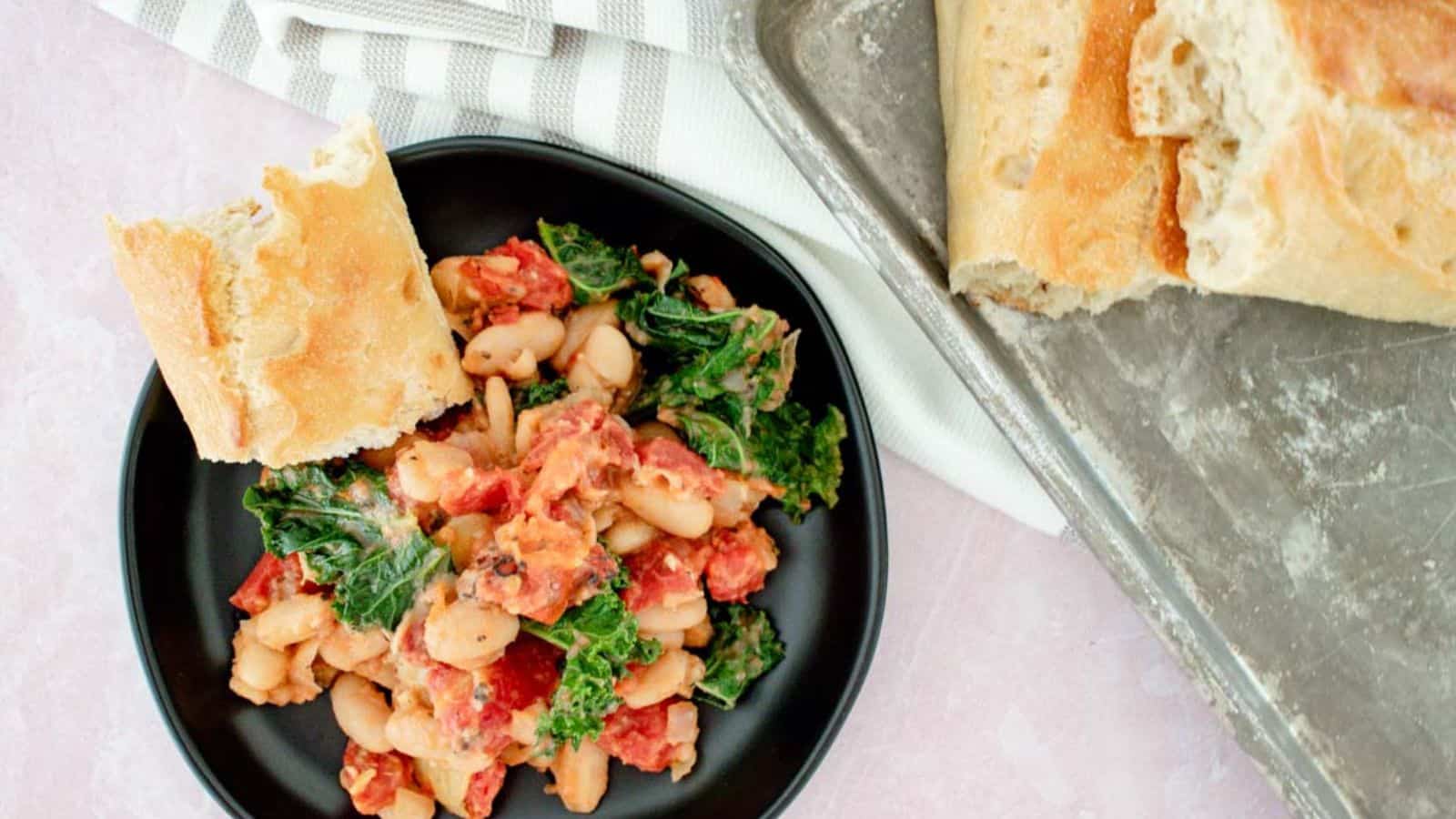 A black plate with white beans, tomatoes, and greens served with a piece of bread. A tray with more bread is visible beside it.