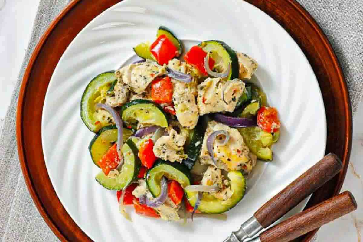 A white plate with a serving of Italian Herbed Chicken & baked foil packs with a fork on the side and a small bowl of seasoning in the background.