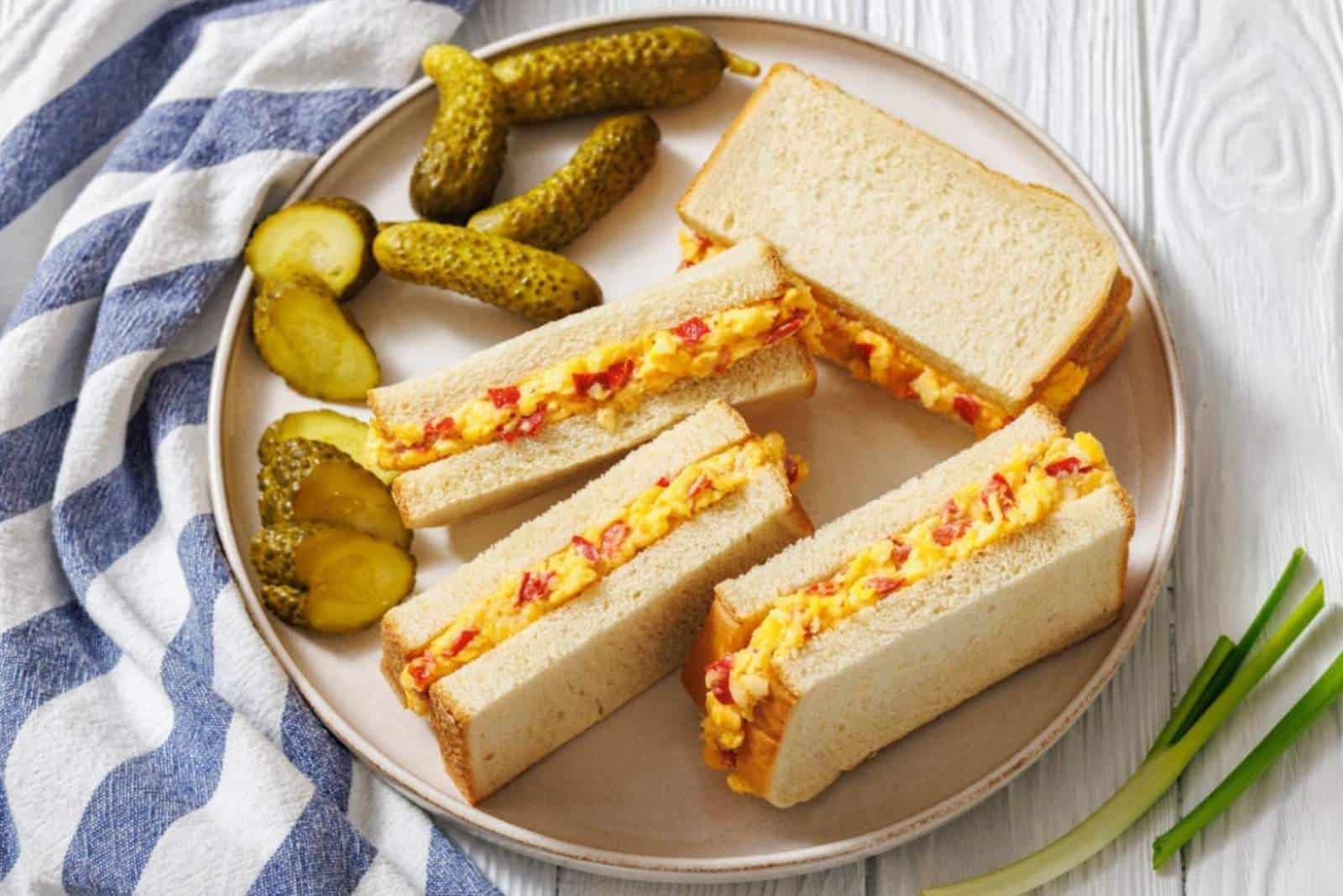 A plate with three pimento cheese sandwiches, potato chips, and pickles, with lettuce and more chips in the background.